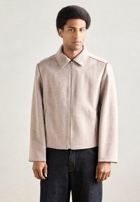 Beżowa wełniana kurtka z kołnierzem, przednim zamkiem błyskawicznym i subtelnymi haftowanymi detalami na kołnierzu. Długie rękawy i krótki krój.