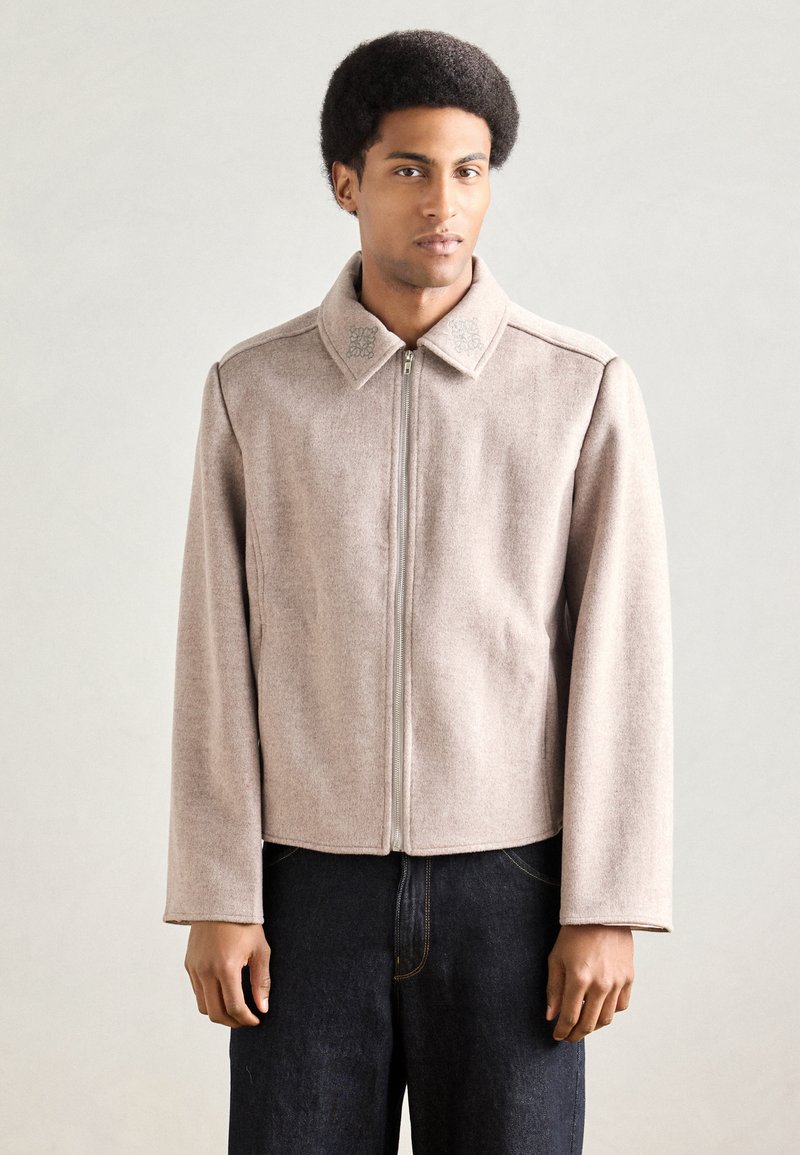 Beżowa wełniana kurtka z kołnierzem, przednim zamkiem błyskawicznym i subtelnymi haftowanymi detalami na kołnierzu. Długie rękawy i krótki krój.