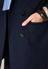 Cappotto navy doppiopetto con bottoni neri, una texture liscia in lana e una tasca laterale. Una mano è visibile mentre è appoggiata nella tasca.