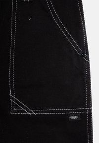 Tejido de mezclilla negra con costuras blancas, que presenta un detalle de bolsillo y una pequeña etiqueta de marca en la parte inferior. Superficie texturizada, diseño robusto.