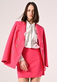 Bright pink tailored blazer with gold buttons over a white blouse featuring a ruffled detail, paired with a matching pink mini skirt.