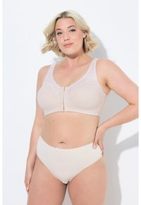 Femme aux cheveux blonds courts portant un soutien-gorge beige à fermeture frontale avec des détails en dentelle et une culotte taille haute assortie, sur fond blanc.