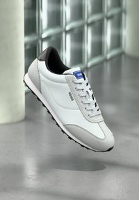 Baskets blanches Hugo avec accents gris et noirs, lacets blancs et logo bleu sur la languette, présentées sur un fond carrelé réfléchissant.