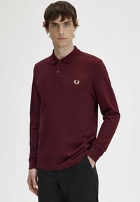 Polo a maniche lunghe color maroon con colletto e tre bottoni, caratterizzato da un logo ricamato bianco sul lato sinistro del petto. Colore uniforme, texture liscia.