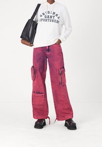 Sudadera blanca con capucha y texto en azul marino, combinada con pantalones cargo de mezclilla rosa de gran tamaño que presentan bolsillos y cremalleras. Zapatos de plataforma negros.