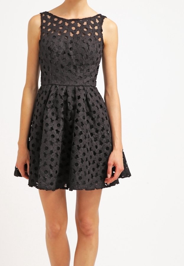 Robe noire sans manches avec superposition en dentelle florale, taille cintrée et jupe évasée, portée par une personne debout devant un fond blanc.