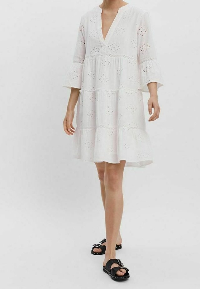 Vit miniklänning med V-ringning, ögla detaljer, utställda ärmar och lager på lager kjol. Bärs med svarta sandaler med dekorativa nitar.