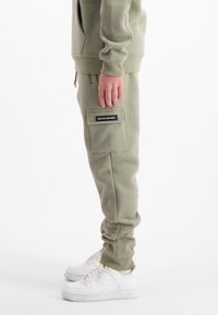 Zijaanzicht van een persoon die licht olijfgroene cargo-broeken draagt met een klepzak en witte sneakers op een witte achtergrond.