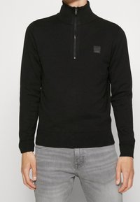 Svart zip-up tröja med hög krage och ribbade muddar. Har en liten fyrkantig logotyp på bröstet och en slät, texturerad tyg.