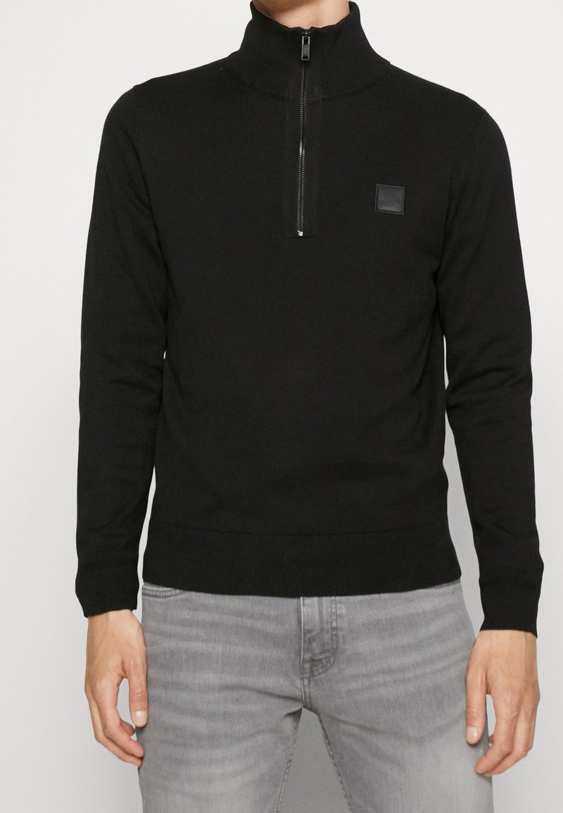 Svart zip-up tröja med hög krage och ribbade muddar. Har en liten fyrkantig logotyp på bröstet och en slät, texturerad tyg.