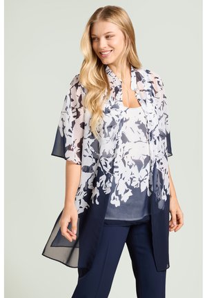 Uśmiechnięta kobieta o długich blond włosach, ubrana w przejrzysty granatowo-biały kwiatowy kimono na pasującym topie i granatowych spodniach.