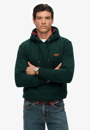 Donkergroene hoodie met trekkoord, voorzien van een geborduurd bruin logo. Gelaagd over een rood en zwart geruit overhemd, gedragen met blauwe spijkerbroek.