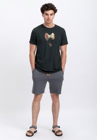 Zwarte T-shirt met een afbeelding van een appelkern, gecombineerd met grijze shorts met gele accenten, en een model draagt bruine sandalen.