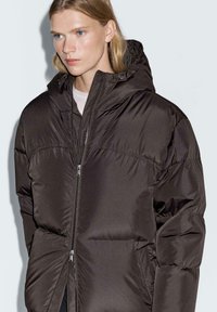 Giovane con lunghi capelli biondi che indossa una giacca piumino oversize marrone scuro con cappuccio e chiusura a doppia zip, sfondo neutro.