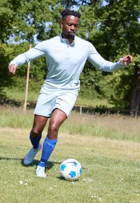 Man in light blue sportswear and blue socks controlling a white and blue soccer ball on grassy field with trees in background.