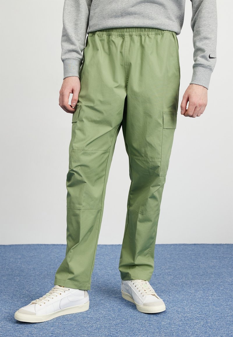 Ljusgröna cargobyxor med elastisk midja, sidofickor och lös passform, parat med vita sneakers på en blå matta.