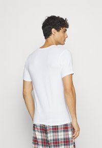 Weißes Baumwoll-T-Shirt mit Rundhalsausschnitt und kurzen Ärmeln, von der Rückseite gezeigt. Kombiniert mit karierten Schlafanzughosen in Rot, Blau und Creme.