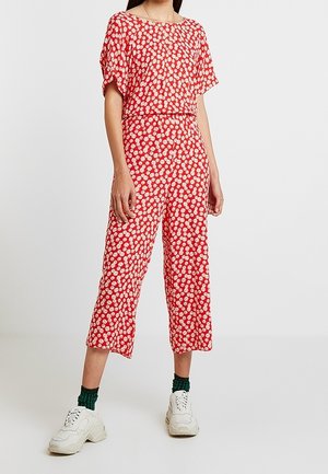 Vrouw draagt een rode bloemenprint korte mouwen jumpsuit met cropped wijde broekspijpen, groene sokken en witte sneakers tegen een effen achtergrond.