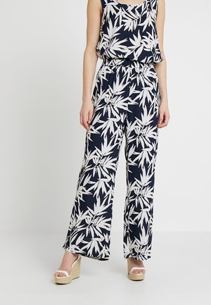 Femme portant un top sans manches bleu marine et blanc à motif feuilles et un pantalon large assorti avec des sandales compensées beige.