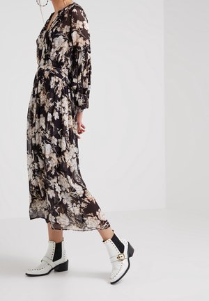 Femme portant une longue robe noire transparente à fleurs et des bottines blanches avec des clous dorés et un détail de boucle sur un fond clair uni.