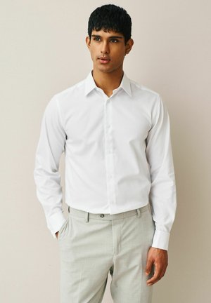 Junger Mann, der ein weißes Hemd mit Knöpfen und hellgraue Hose trägt, steht mit einer Hand in der Tasche vor einem schlichten beigefarbenen Hintergrund.