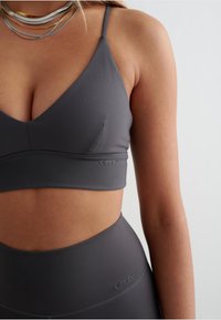 Gray athletic bra with a V-neck design, made of smooth fabric. Features thin straps and a subtle logo on the band, paired with matching high-waisted leggings.