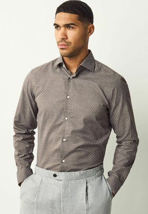 Cămașă lungă cu mânecă, maro, cu un model de buline albe, cu guler cu nasturi și nasturi pe față. Purtați-o cu pantaloni gri deschis, plisati.