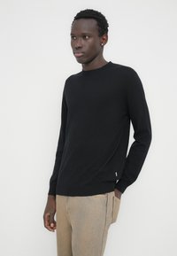 Pull en maille noir avec col rond, manches longues et ourlet côtelé. Comprend une petite étiquette de marque sur le côté. Porté avec un jean beige.
