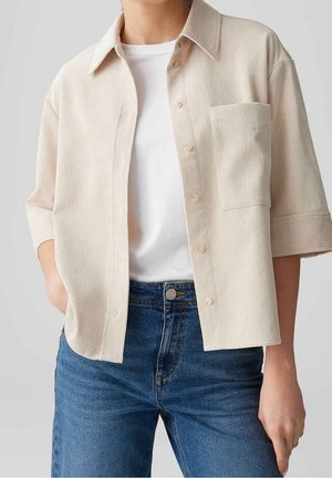 Persoon draagt een wit T-shirt onder een beige overhemd met korte mouwen, knopen en een borstzak, en blauwe spijkerbroek.