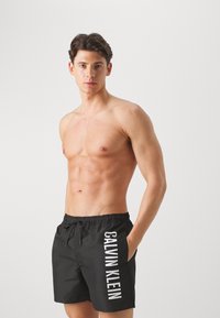 Crne kupaćice kratke hlače s elastičnom vrpcom i džepovima, koje imaju istaknuti bijeli logo "CALVIN KLEIN" duž bočne strane.