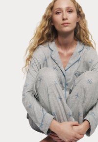 Grå pyjamasset med ljusblå accenter, bestående av en knäppningstopp och byxor, dekorerad med små blå rosettmönster. Mjuk, lätt vikt tyg.