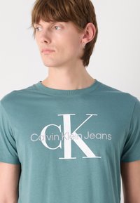 Pashipinta turkio spalvos trumpas rankovėmis marškinėliai, pagaminti iš minkštos medvilnės, su dideliu baltu "CK" ženkleliu ir šviesiai pilku šriftu užrašu "Calvin Klein Jeans".