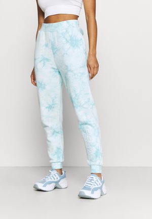 Vrouw draagt lichtblauwe en witte tie-dye joggers met de tekst "GUESS" op het been, een wit cropped topje en lichtblauwe en witte sneakers, staand.