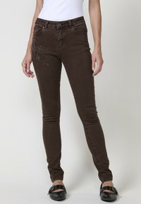 Los jeans skinny de mezclilla marrón presentan un patrón floral con acentos metálicos en el muslo izquierdo y bolsillos funcionales. La cinturilla tiene un cierre de botón.