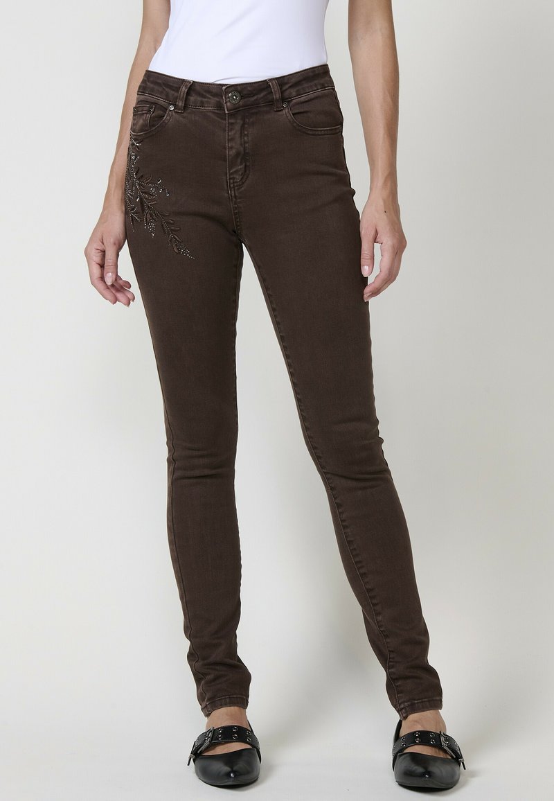 Los jeans skinny de mezclilla marrón presentan un patrón floral con acentos metálicos en el muslo izquierdo y bolsillos funcionales. La cinturilla tiene un cierre de botón.