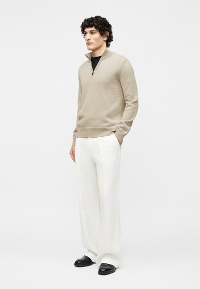 Noor mees seisab, seljas beež toonis neljandiku lukuga kampsun, must särgike, valged laia säärega püksid ja mustad jalanõud, valgel taustal.