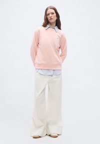 Γυναίκα στέκεται φορώντας ανοιχτό ροζ φούτερ πάνω από μπλε πουκάμισο με γιακά, φαρδιά λευκά παντελόνια και μπεζ παπούτσια σε απλό φόντο.