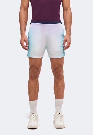 Uomo che indossa pantaloncini sportivi con sfumature bianco e blu, calzini bianchi, scarpe da ginnastica bianche e una maglietta viola, in piedi con le braccia rilassate lungo i fianchi.