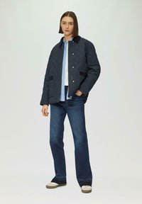 Veste noire matelassée avec col, fermeture à pression sur le devant et poches latérales ; associée à un jean bleu foncé et des baskets bicolores.