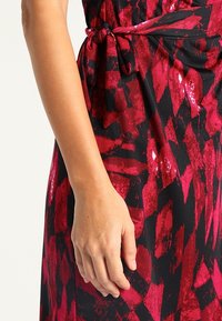 Punainen ja musta kuviollinen mekko, jossa on sidottu vyötärö. Kankaan pinnassa on paljettitekstuuri ja geometrinen muotoilu, korostaen eläviä värejä.