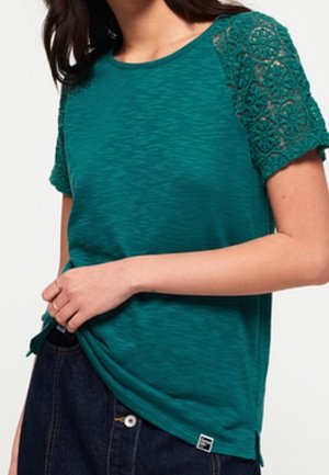 Vrouw die een zeegroen T-shirt draagt met korte kanten mouwen en donkere spijkerbroek, handen passen de zoom van het shirt aan.