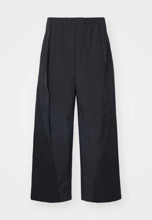 Schwarze weit geschnittene Hose mit einem elastischen Bund, seitlichen Reißverschlusstaschen und schrägen Nähten für zusätzliche Struktur. Hergestellt aus einem glatten, leichten Stoff.