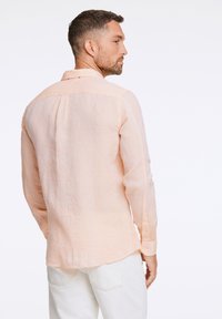 Camisa de lino color durazno claro con mangas largas, cuello abotonado y una textura sutil. Presenta un corte recto y detalles de pliegue en la parte posterior.