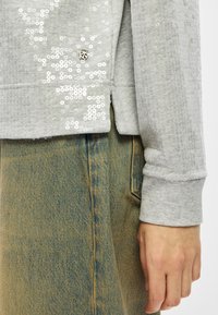 Mão descansando ao lado, vestindo jeans azuis desbotados e um suéter cinza com lantejoulas e o logotipo da DKNY perto da barra.