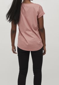 Pink short-sleeve shirt with rolled cuffs, rounded hem, and soft fabric. Worn with black fitted pants. Rear view shown.