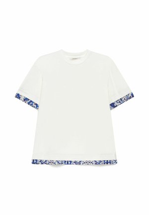 Camicia bianca a maniche corte con bordo a motivo floreale blu e bianco sui bordi delle maniche e sull'orlo inferiore, stesa in piano su sfondo bianco.