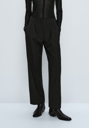 Personne portant un pantalon noir ample taille haute avec des poches, associé à des bottines noires à bout pointu et un haut noir ajusté.