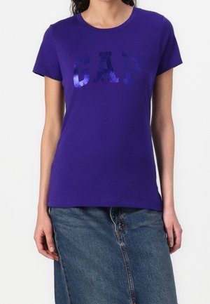Femme portant un T-shirt violet à manches courtes avec un logo "GAP" brillant, associé à une jupe en jean bleue, debout devant un fond blanc.
