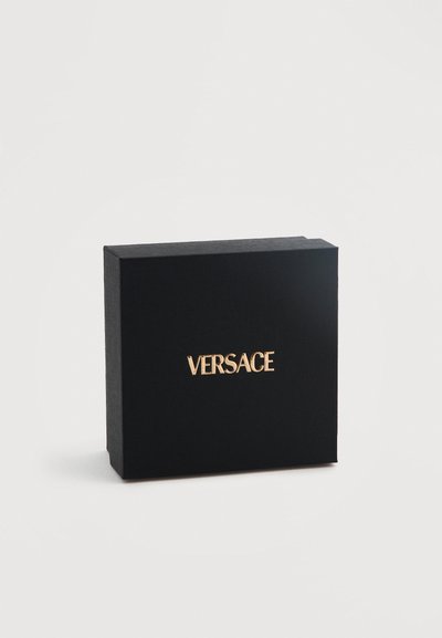 Μαύρο ορθογώνιο κουτί με υφή στην επιφάνεια και χρυσό ανάγλυφο λογότυπο "VERSACE" στο κέντρο της μπροστινής πλευράς, σε λευκό φόντο.