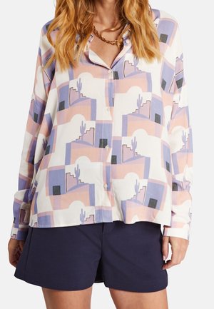 Vrouw draagt een pastel gekleurd overhemd met geometrische patronen en cactussen, gecombineerd met donkerblauwe shorts en een gouden ketting.